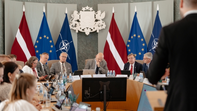 Ministru kabineta sēžu zālē ap apaļu galdu sēž vīrieši un sievietes, kas piedalās diskusijā un strādā ar datoriem un dokumentiem. Telpas centrā redzams Latvijas valsts ģerbonis, bet fonā izvietoti Latvijas, Eiropas Savienības un NATO karogi. Priekšplānā redzama stāvoša persona, kas uzrunā sanāksmes dalībniekus.
