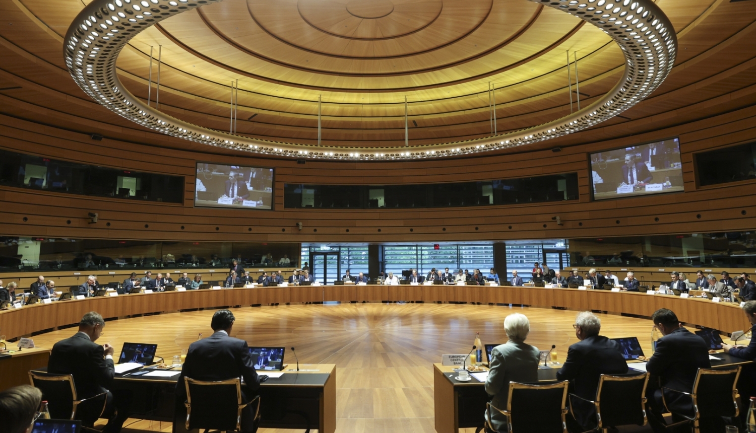 Apļveida konferenču zāle, kurā aplī izkārtots galds, pie kura sēž sievietes un vīrieši, pie sienām ir projektora ekrāni, virs galvām - apaļa lampa