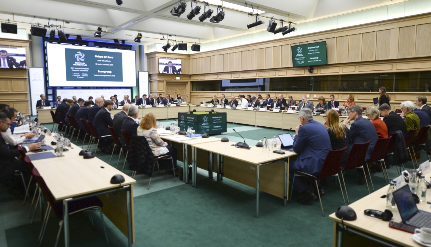 Lielā konferenču zālē izkārtoti galdi, pie kuriem sēž cilvēki, uz galdiem stāv dokumenti, portatīvie datori un mikrofoni