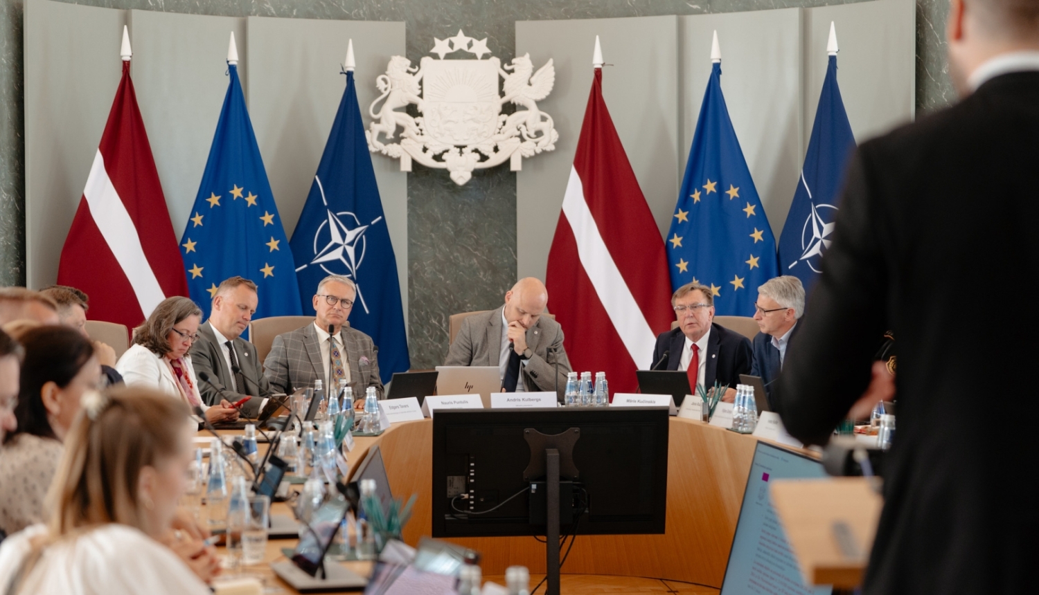 Ministru kabineta sēžu zālē ap apaļu galdu sēž vīrieši un sievietes, kas piedalās diskusijā un strādā ar datoriem un dokumentiem. Telpas centrā redzams Latvijas valsts ģerbonis, bet fonā izvietoti Latvijas, Eiropas Savienības un NATO karogi. Priekšplānā redzama stāvoša persona, kas uzrunā sanāksmes dalībniekus.