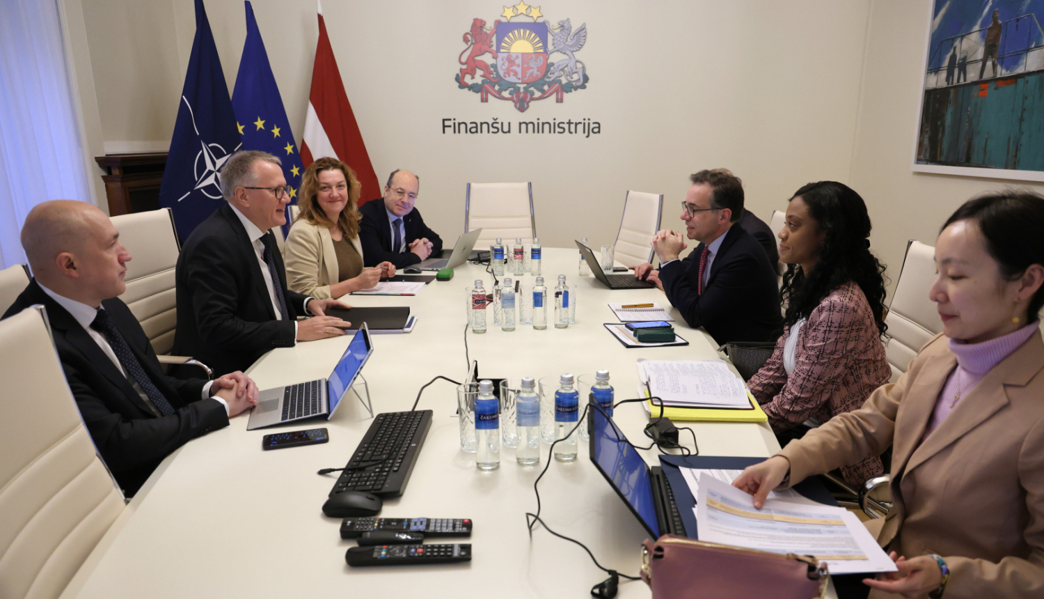 Attēlā redzama sanāksmju telpa Finanšu ministrijā: ap galdu sēž ministrijas pārstāvji un delegācijas locekļi, uz galda redzami klēpjdatori un dokumenti, fonā redzami NATO, ES un Latvijas karogi un uz sienas uzraksts “Finanšu ministrija”
