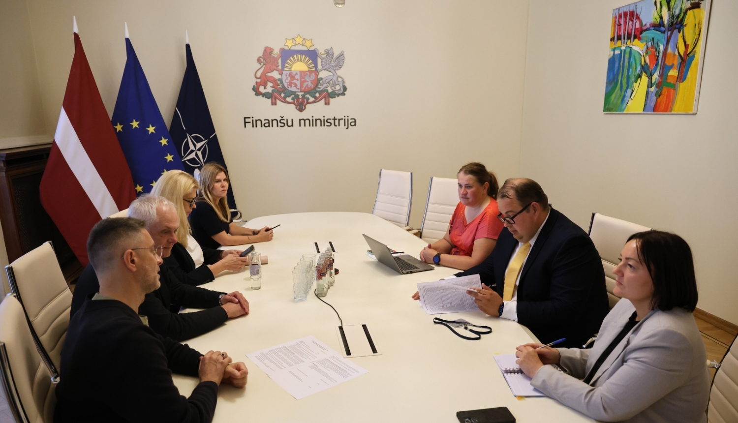 Gaišā telpā pie ovāla galda sēž sievietes un vīrieši, savstarpēji diskutē, uz gada stāv dokumenti, telpas stūrī redzami Latvijas, Eiropas Savienības un NATO karogi, uz sienas ir Finanšu ministrijas ģerbonis