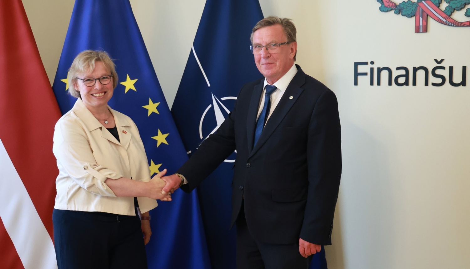 Finanšu ministrs Māris Kučinskis ar Vācijas Federatīvās Republikas ārkārtējo un pilnvaroto vēstnieci Gudrunu Maslohu stāv telpā un sarokojas. Fonā redzami Latvijas, Eiropas Savienības un NATO karogi.