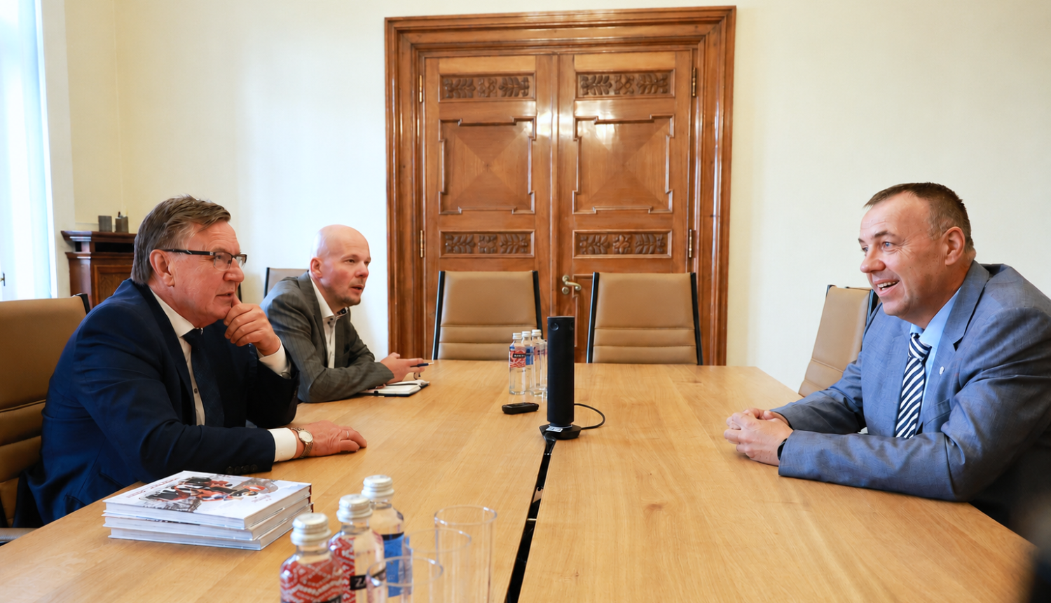 Darba tikšanās sanāksmju telpā, kurā trīs vīrieši uzvalkos apspriež jautājumus pie konferenču galda