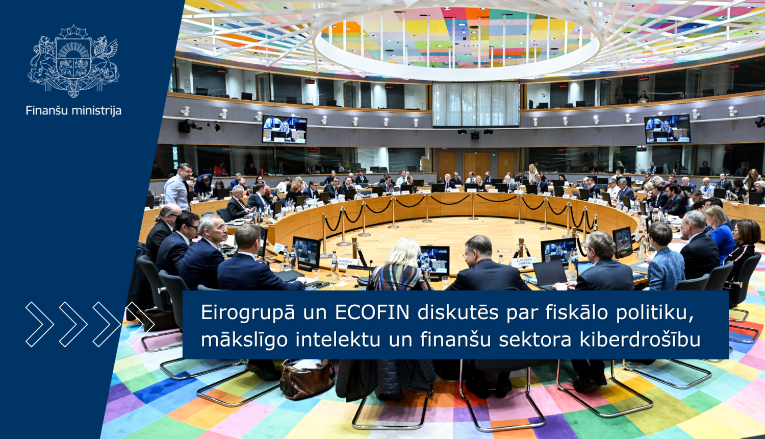 Plašā konferenču zālē ar krāsainiem griestiem pie apaļa galda sēž vīrieši un sievietes kostīmos, savstarpēji diskutē