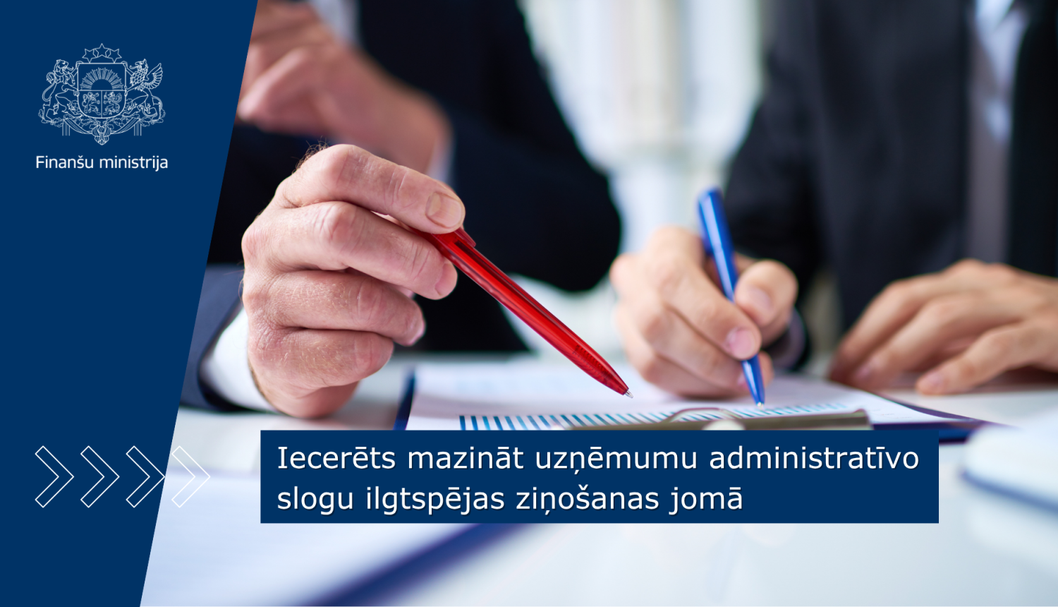 Divi vīrieši sēž pie gada un veic labojumus uz galda esošos dokumentos, vīriešiem rokās redzamas sarkana un zila pildspalva 