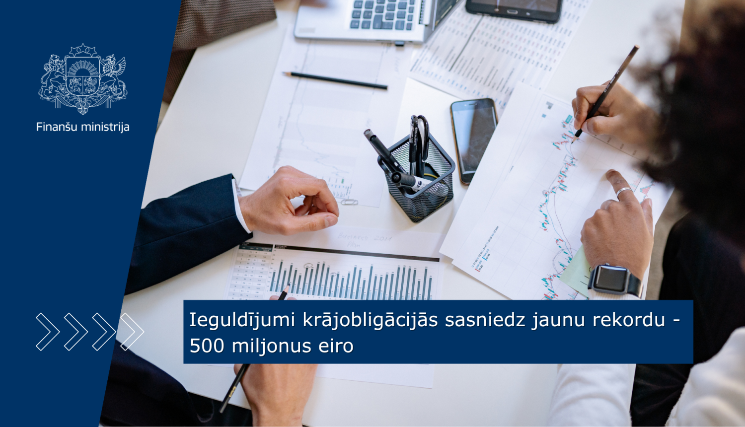 Uz galda stāv dokumenti ar dažādiem grafikiem un datiem, divi cilvēki tos pēta, veicot piezīmēs tajos