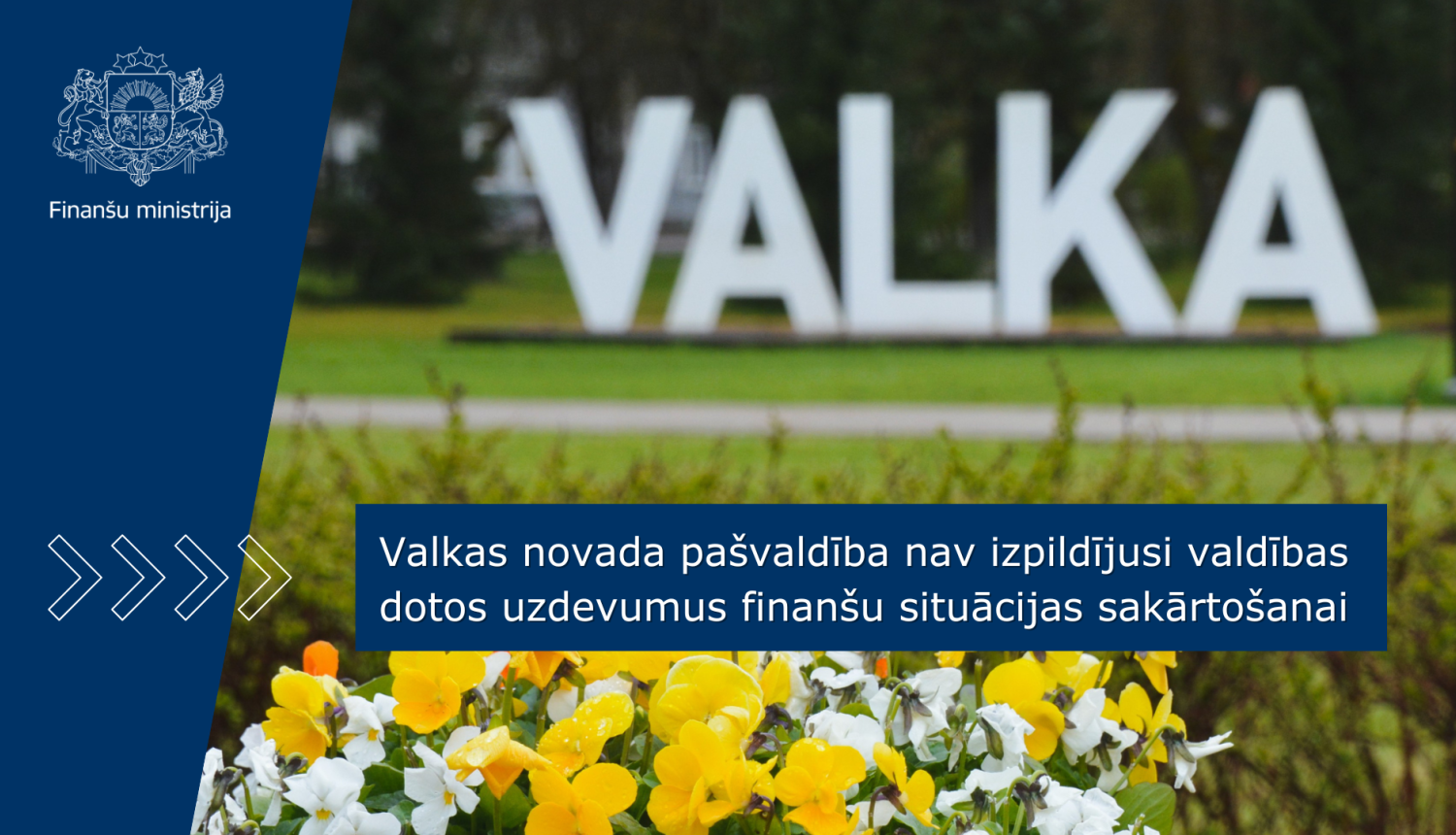 Dabā stāvošs VALKAS pilsētas uzraksts - burti, priekšplānā ir redzami ziedi. Uz attēla ir teksts