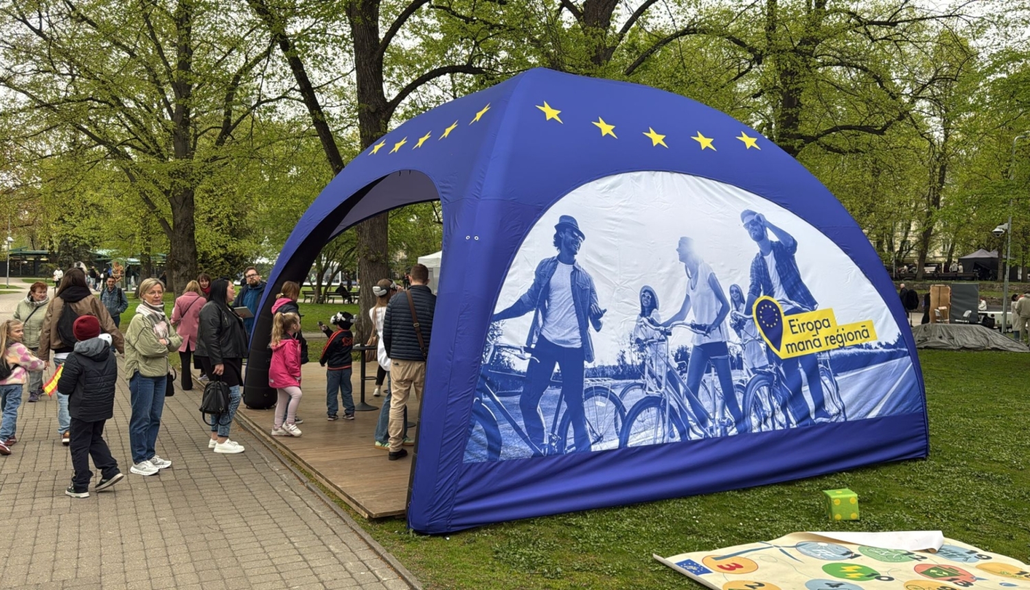 Apdrukāta telts parkā, cilvēki iet iekšā un nāk ārā no telts