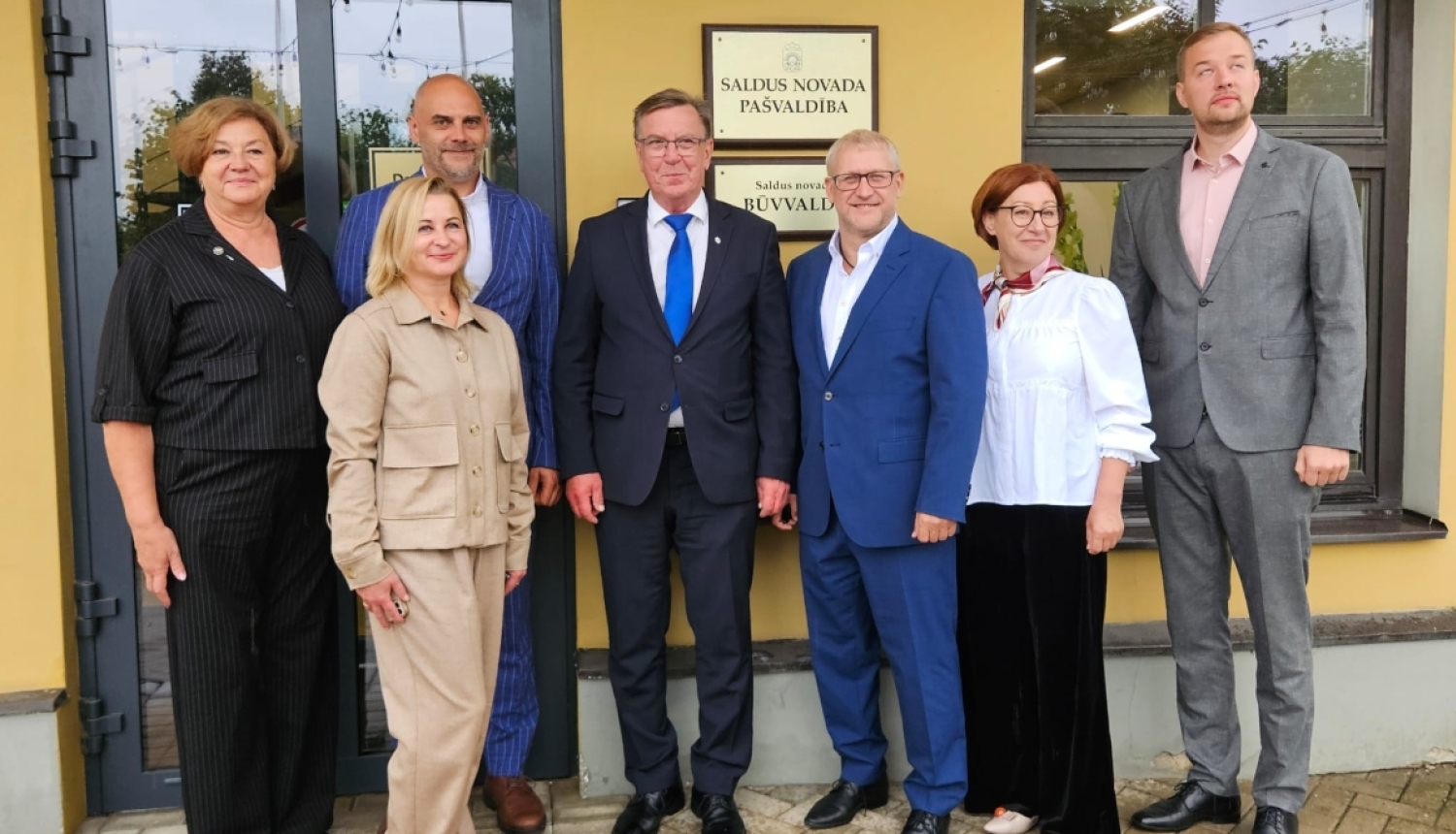 Pie ēkas ieejas stāv sievietes un vīrieši un skatās kameras objektīvā