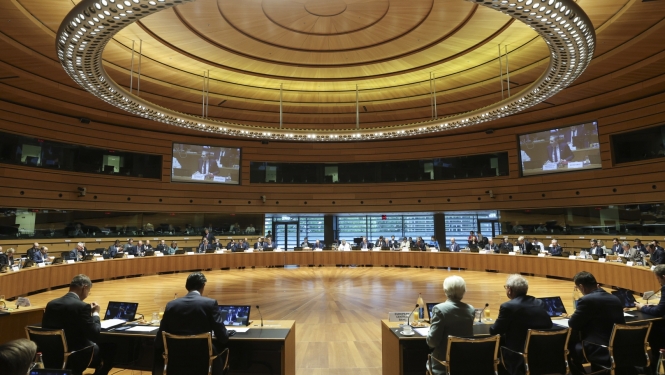 Apļveida konferenču zāle, kurā aplī izkārtots galds, pie kura sēž sievietes un vīrieši, pie sienām ir projektora ekrāni, virs galvām - apaļa lampa