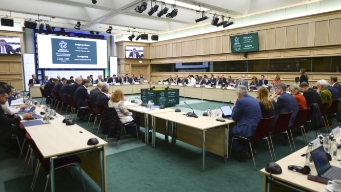 Lielā konferenču zālē izkārtoti galdi, pie kuriem sēž cilvēki, uz galdiem stāv dokumenti, portatīvie datori un mikrofoni
