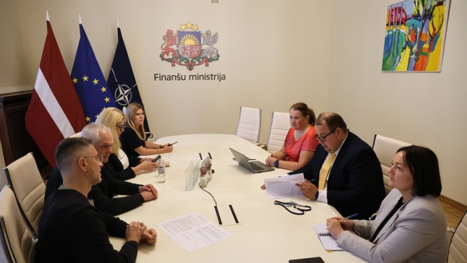 Gaišā telpā pie ovāla galda sēž sievietes un vīrieši, savstarpēji diskutē, uz gada stāv dokumenti, telpas stūrī redzami Latvijas, Eiropas Savienības un NATO karogi, uz sienas ir Finanšu ministrijas ģerbonis
