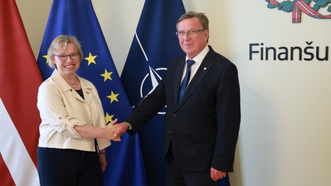 Finanšu ministrs Māris Kučinskis ar Vācijas Federatīvās Republikas ārkārtējo un pilnvaroto vēstnieci Gudrunu Maslohu stāv telpā un sarokojas. Fonā redzami Latvijas, Eiropas Savienības un NATO karogi.
