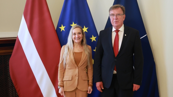 Vīrietis un sieviete stāv, vērsti pret kameru, aiz muguras ir Latvijas, Eiropas Savienības un Nato karogi