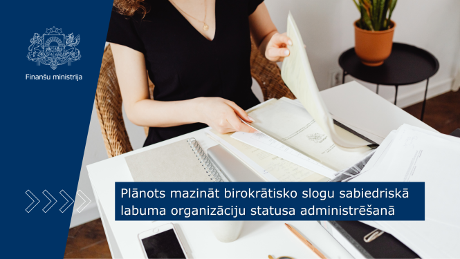 Pie galda sēž sieviete un strādā ar dokumentiem, uz galda stāv arī portatīvais dators, telefons un pierakstu klad