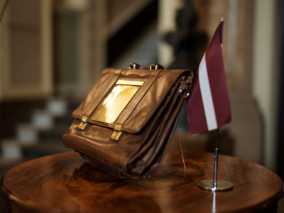 Attēlā redzams tradicionālais budžeta portfelis statīvā novietots uz apaļa galdiņa, līdzās stāv Latvijas karodziņš