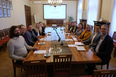 Attēlā pie gara galda abpus galdam sēž vīrieši un sievietes un smaidot skatās kamerā, tālumā pie sienas ir redzams projektora ekrāns