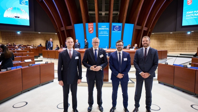 Attēlā redzami četri vīrieši uzvalkos, kuri pozē oficiālā vidē – konferenču zālē