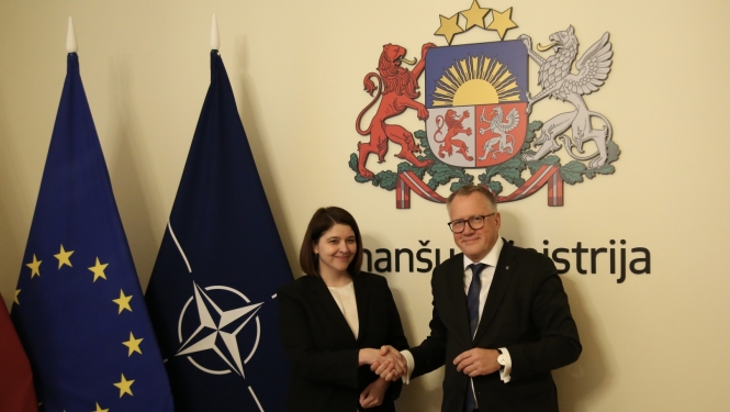 Sieviete melnā klasiska kostīmā sarokojas ar vīrieti uzvalkā stāvot pie karogiem