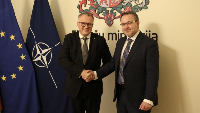 Divi vīrieši tumšos uzvalkos sarokojas uz ES un NATO karogu un Finanšu ministrijas ģērboņa fona
