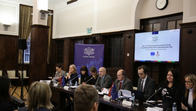 Rindā pie galda sēž vairāki cilvēki, fornā redzami TV ekrān ar tekstveida informāciju