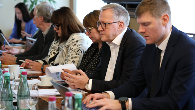 Formāli ģērbušos cilvēku grupa sēž pie galda un piedalās sanāksmē. Daži strādā ar datoru, citi pēta dokumentus. Uz galda stāv glāzes un ūdens pudeles