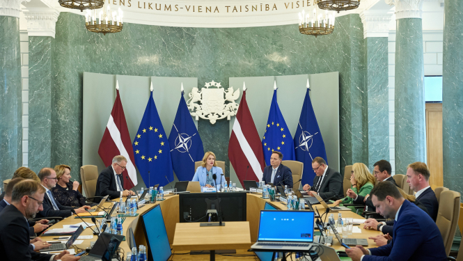 Attēlā redzama Ministru kabineta sēde, kurā visi ministri un Ministru prezidenta sēž pie galda un savstarpēji diskutē, telpas galā pie sienas stāv karogi
