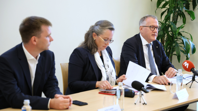 Preses konference. Trīs cilvēki sēž pie galda, uz tā novietoti mikrofoni, dokumenti, ūdens glāzes.