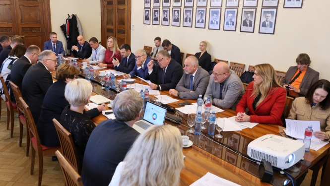 Attēlā garš galds ap kuru no visām pusēm sēž vīrieši un sievietes un savstarpēji sarunājas, uz galda stāv dokumenti, portatīvie datori, ūdens pudeles un glāzes