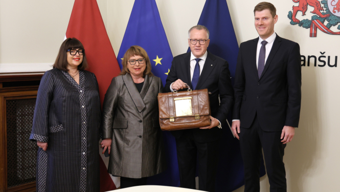 Attēlā uz Latvijas, ES un Nato karogu fona stāv četri cilvēki un smaida, divi no tiem rokās tur portfeli