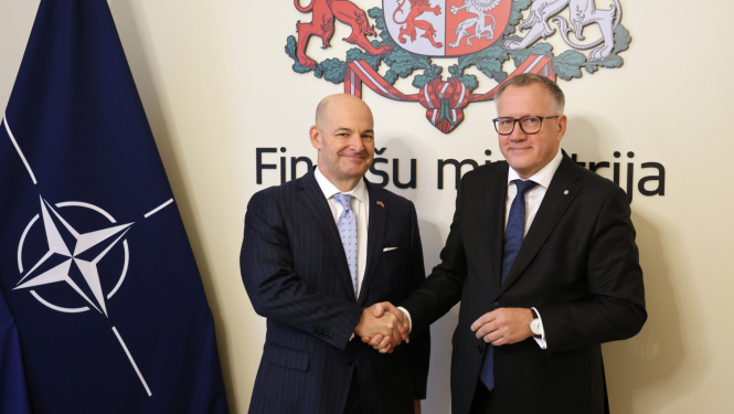 Attēlā uz Finanšu ministrijas ģerboņa fona, sarokojas divi vīrieši uzvalkos, blakus redzams NATO karogs