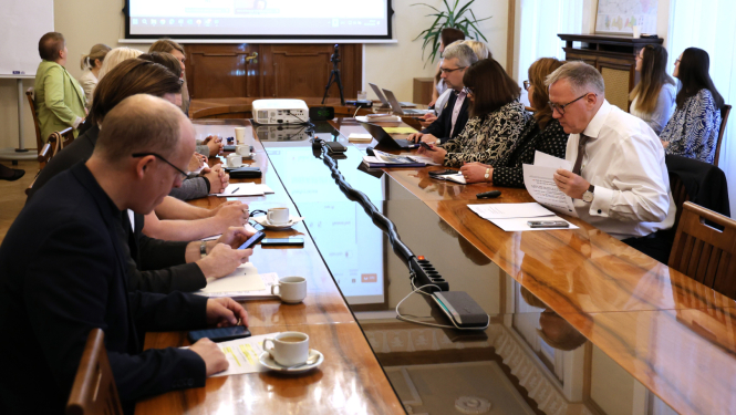 Sanāksmes laikā cilvēki sēž pie gara koka galda, skatās dokumentus, lieto datorus; fonā redzams projekcijas ekrāns ar prezentāciju
