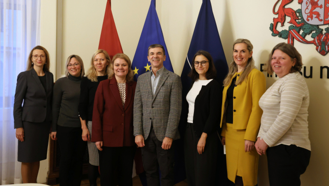 Attēlā uz karogu fona fotografējas rindā stāvošas sievietes un vīrietis, visi smaida