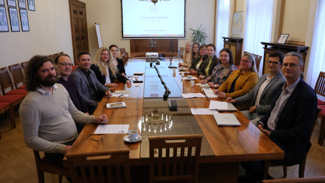 Attēlā pie gara galda abpus galdam sēž vīrieši un sievietes un smaidot skatās kamerā, tālumā pie sienas ir redzams projektora ekrāns