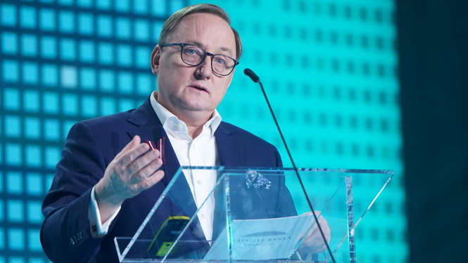 Attēlā tuvplānā redzams vīrietis, kurš stāv pie caurspīdīgas tribīnes un žestikulējot runā, viņas priekšā ir mikrofons