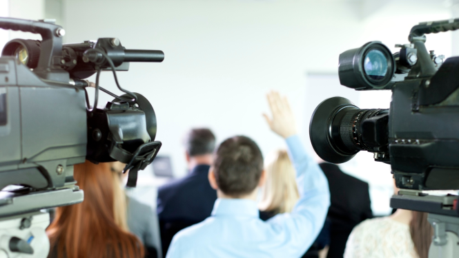 Preses konference ar televīzijas kamerām priekšplānā; auditorijā cilvēks paceļ roku, lai uzdotu jautājumu.