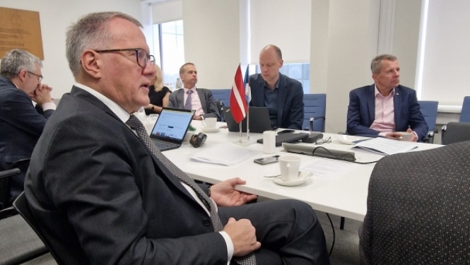 Konferenču zālē pie galda sēž vīrieši uzvalkos un diskutē