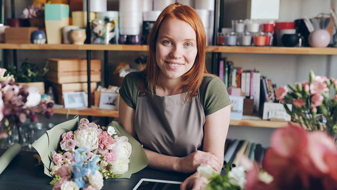 Attēlā redzama rudmate ziedu veikalā pieturot planšetdatoru, kas atrodas uz galda