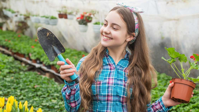 Jauna meitene siltumnīcā tur rokās lāpstiņu un puķupodu