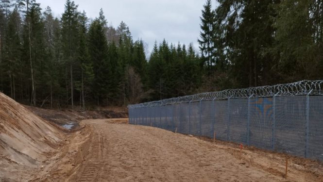 smilšains ceļš mežā, gar kuru redzams metāla žogs ar dzeloņstieplēm