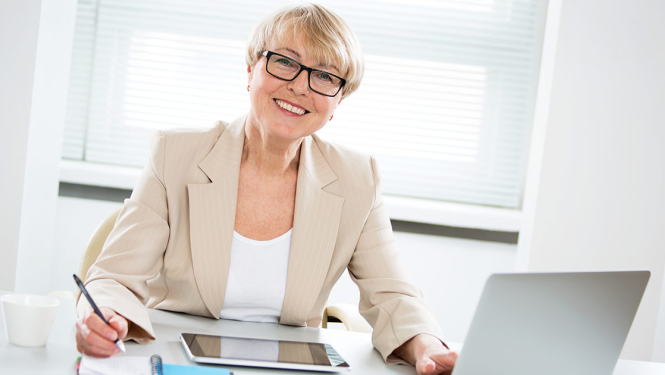 Smaidīga sieviete ar brillēm strādā pie datora.