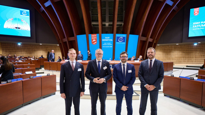 Attēlā redzami četri vīrieši uzvalkos, kuri pozē oficiālā vidē – konferenču zālē