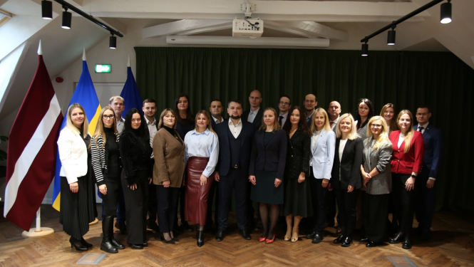 Divās rindās stāv vīrieši un sievietes, fonā Latvijas, Ukrainas un Eiropas Savienības karogi