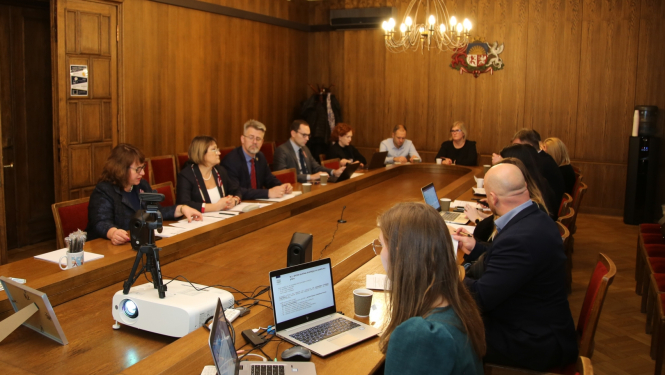 Ap ovālu sanāksmju galdu sēž sievietes un vīrieši