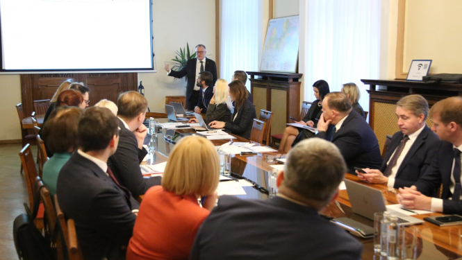 Sēžu zālē pie galda sēž sievietes un vīrieši, galda galā stāvus runā vīrietis