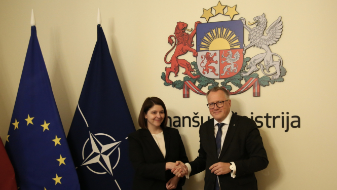 Sieviete melnā klasiska kostīmā sarokojas ar vīrieti uzvalkā stāvot pie karogiem