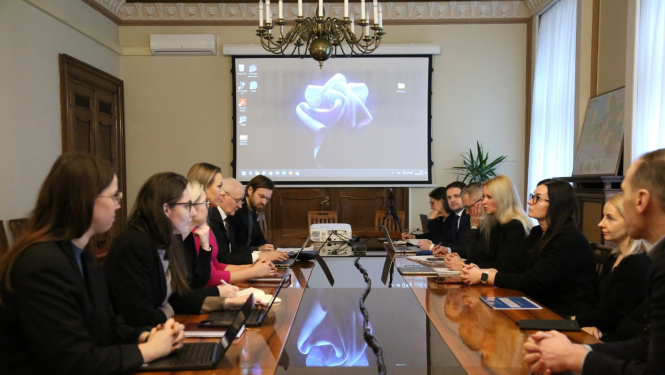 Telpā pie gara galda no abām pusēm sēž sievietes un vīrieši un sarunājas