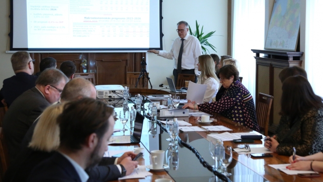 Ap galdu sēž sievietes un virieši un klausās galda galā stāvošā runātāja teiktajā