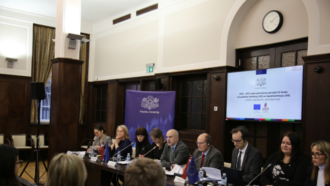 Rindā pie galda sēž vairāki cilvēki, fornā redzami TV ekrān ar tekstveida informāciju